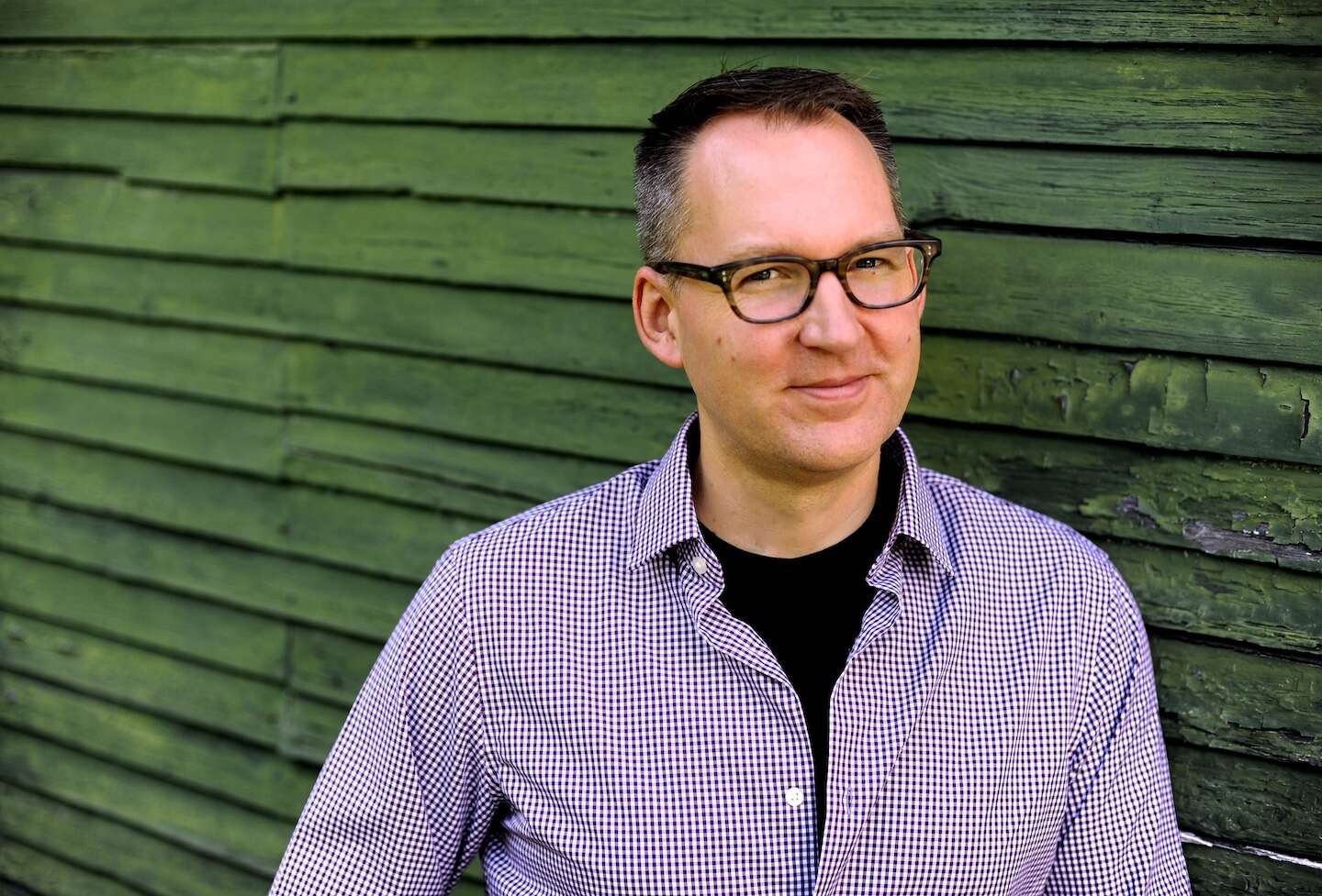A person wearing a blue and white shirt and glasses with a warm grin, standing in front of a green wooden wall.