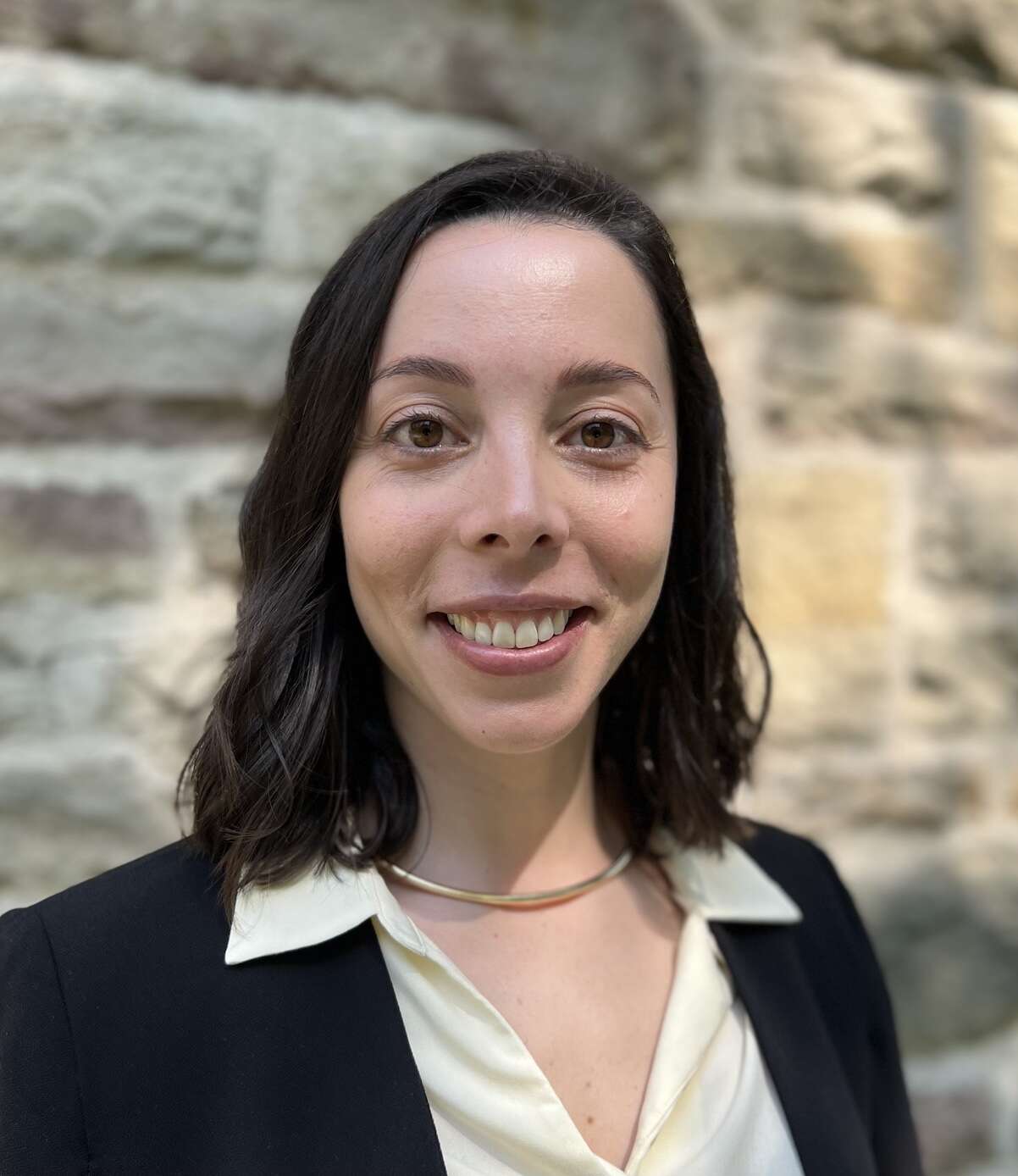 A headshot of a person wearing a white blouse and black blazer, with a gold necklace, smiling in front of a stone wall.