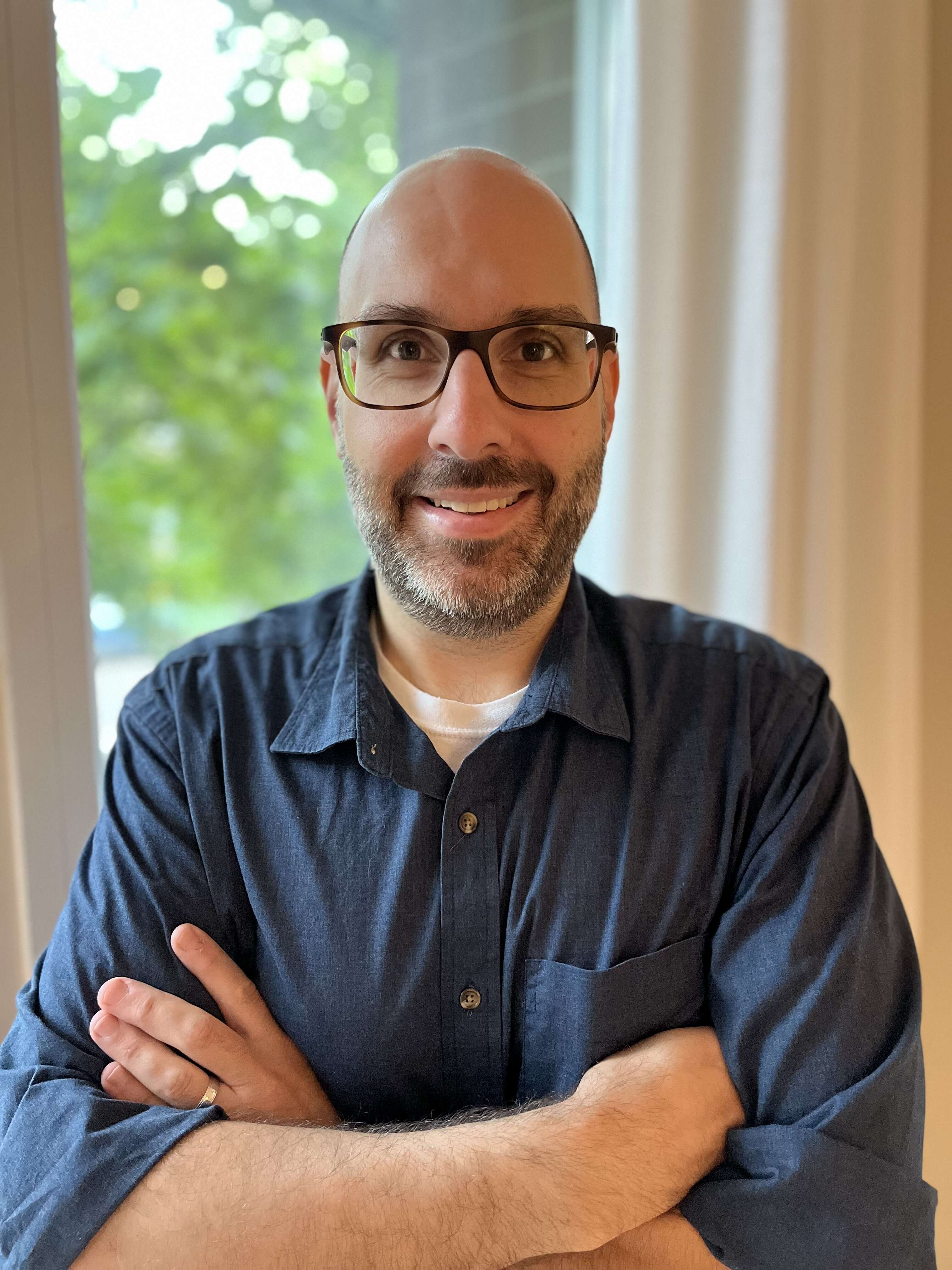 Person with glasses smiling, standing with their arms crossed in front of a window
