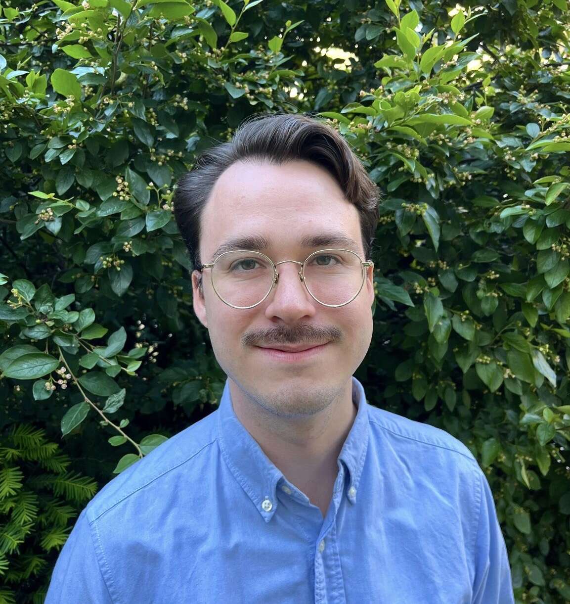 Person wearing glasses and a blue shirt, smiling, in front of trees