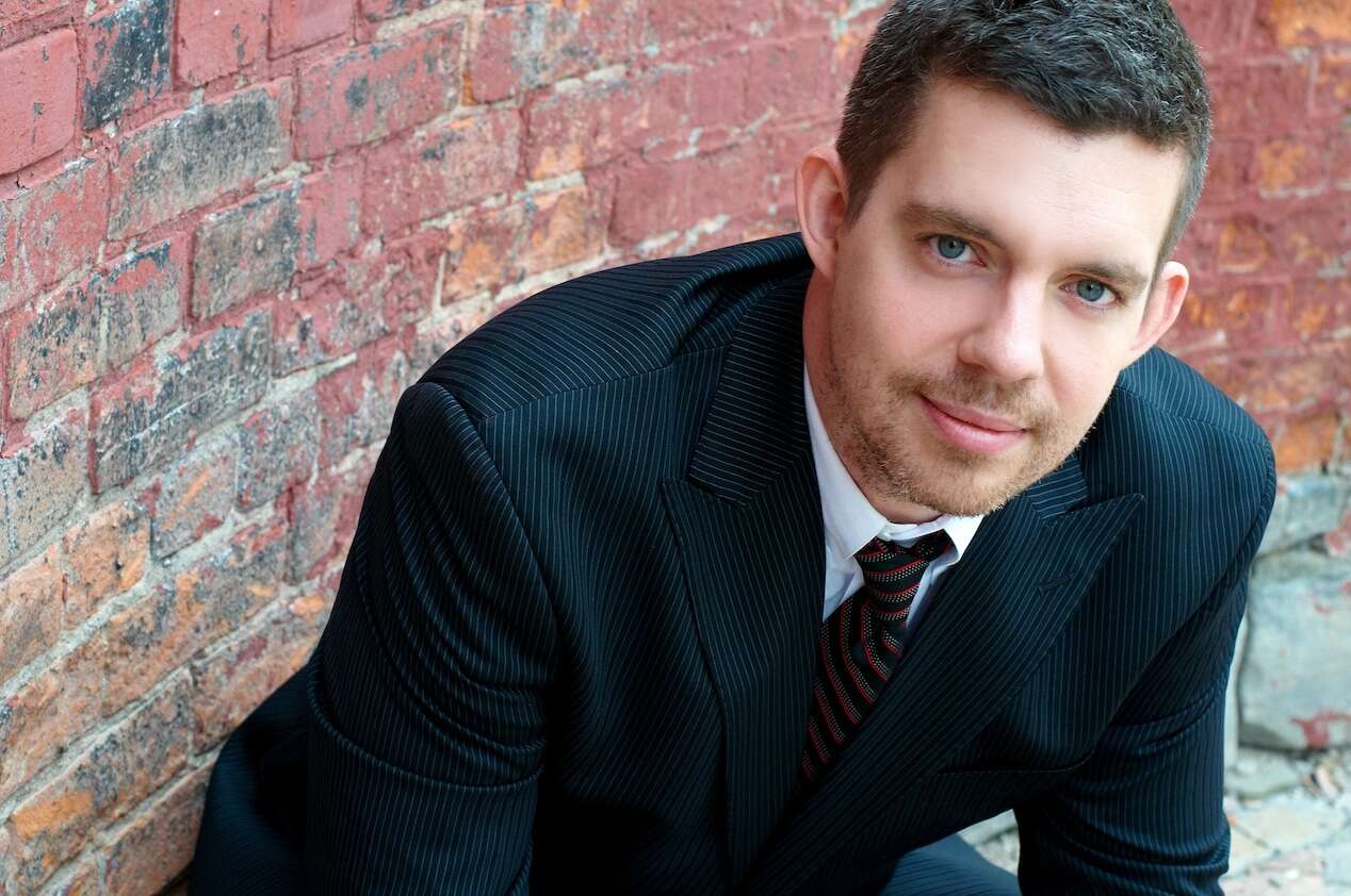 A person in a black suit sitting on a stone surface in front of a brick wall, looking up at the camera.