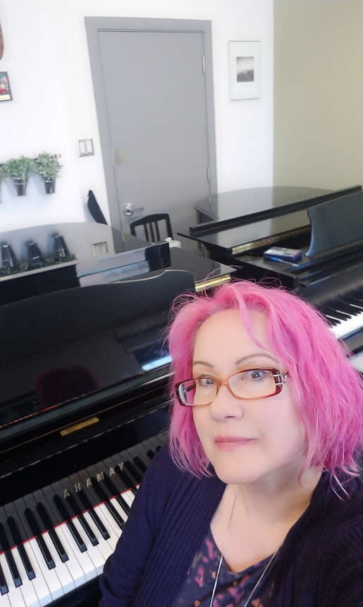 Person with pink hair and glasses posing in front of two grand pianos.