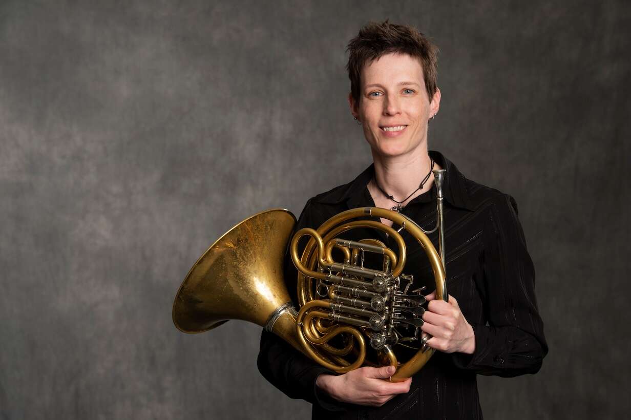 Person smiling, wearing a black shirt, holding a French horn in front of a grey background, creating a balanced and professional appearance.