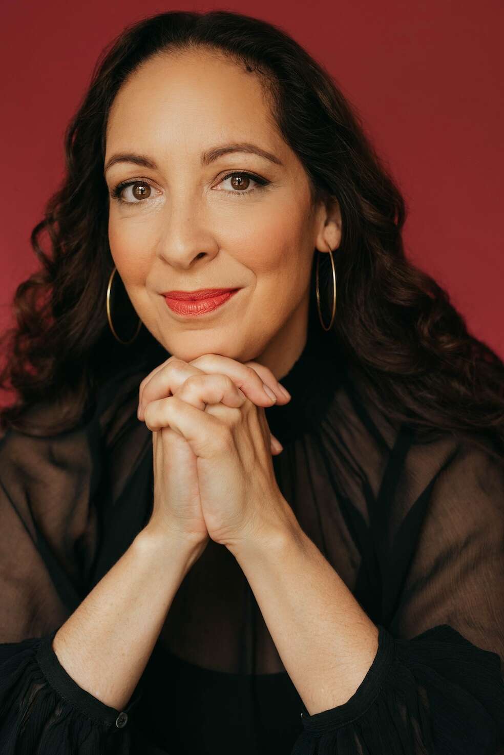Headshot of a person wearing a black shirt and large hoop earrings, resting their head on their hand, with a bold red background adding contrast.