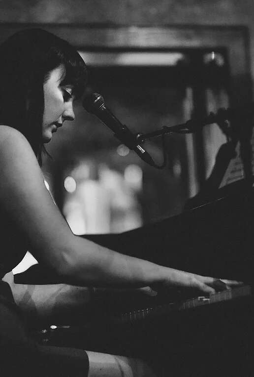 Black and white photo of a person playing the piano and singing with a microphone, with their attention focused on the keys.