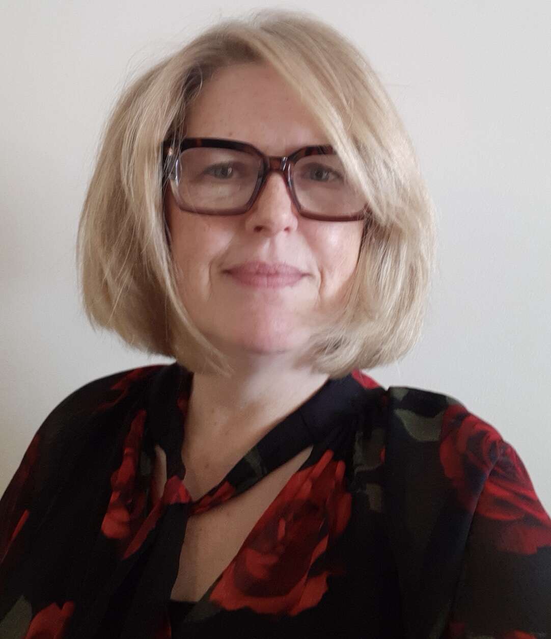 eadshot of a person with glasses and jaw-length blonde hair, wearing a black shirt with a rose pattern, with a warm and approachable expression.