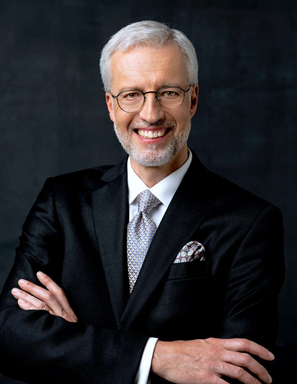 Person smiling, wearing a suit, standing in front of a dark background, exuding confidence and professionalism.
