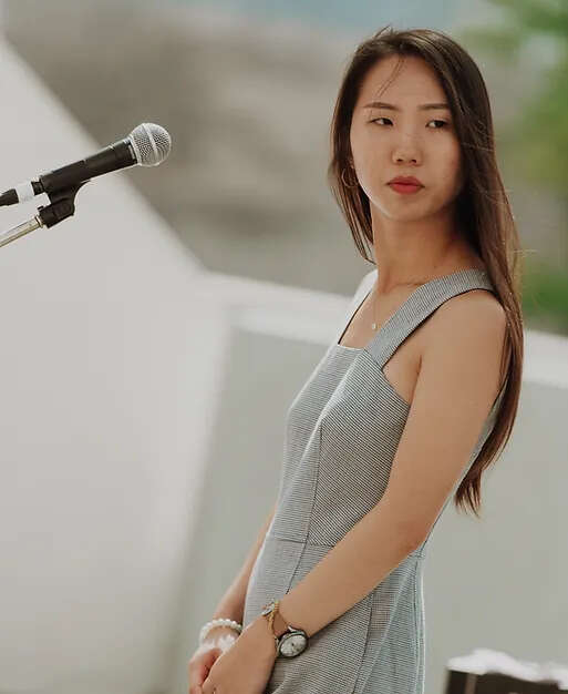 Person in a grey dress standing in front of a microphone, looking toward the side stage, possibly awaiting a cue or preparing for their next move.