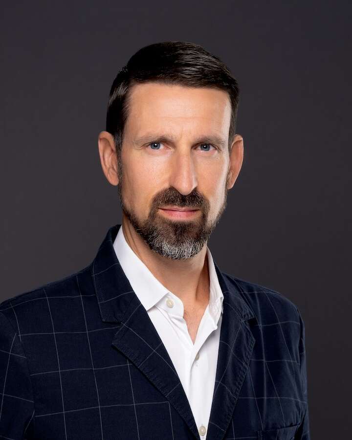 Headshot of a person wearing a blue suit, looking at the camera with a confident expression against a grey background.