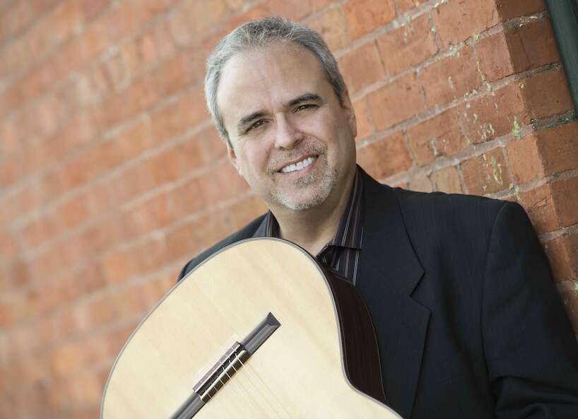 Person wearing a suit, holding a guitar and smiling, standing in front of a brick wall.