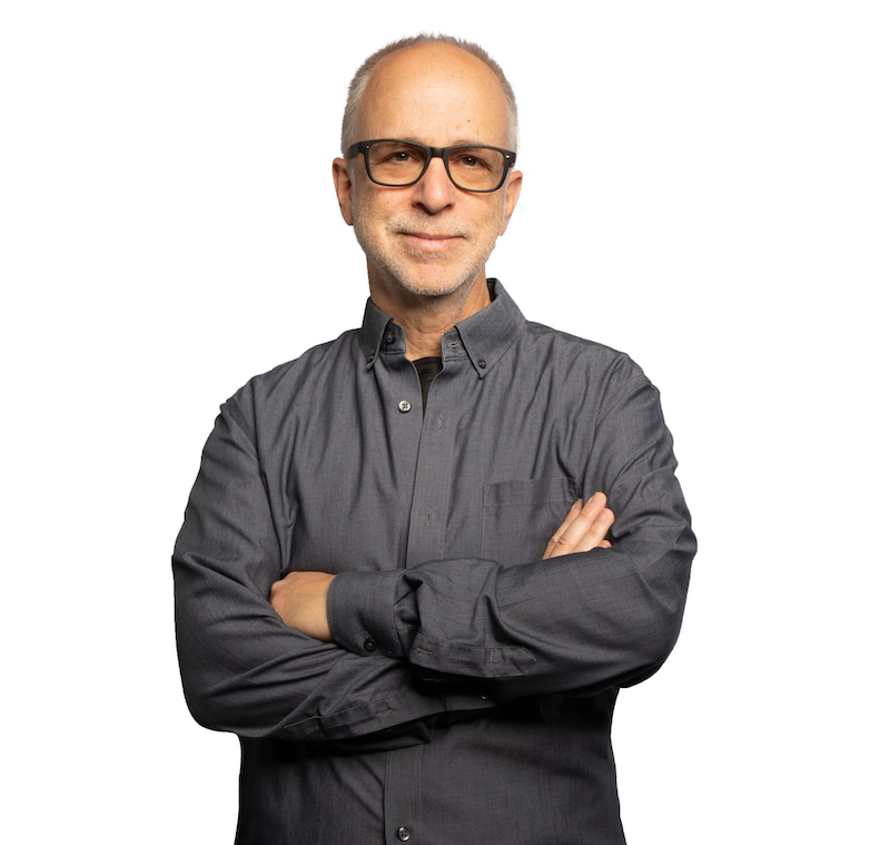 Man wearing glasses and a gray button-down shirt, standing in front of a white backdrop.