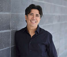Person wearing a black button-down shirt, smiling in front of a grey brick wall.