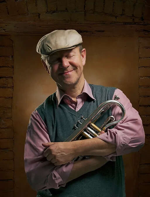 Person wearing a pink shirt and sweater vest, cradling a trumpet in front of a brick wall.