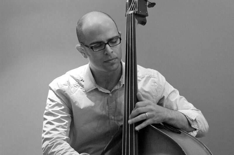 Black and white photo of a person wearing glasses and a button-up shirt, with their eyes closed, playing the double bass, capturing a moment of deep concentration and passion.