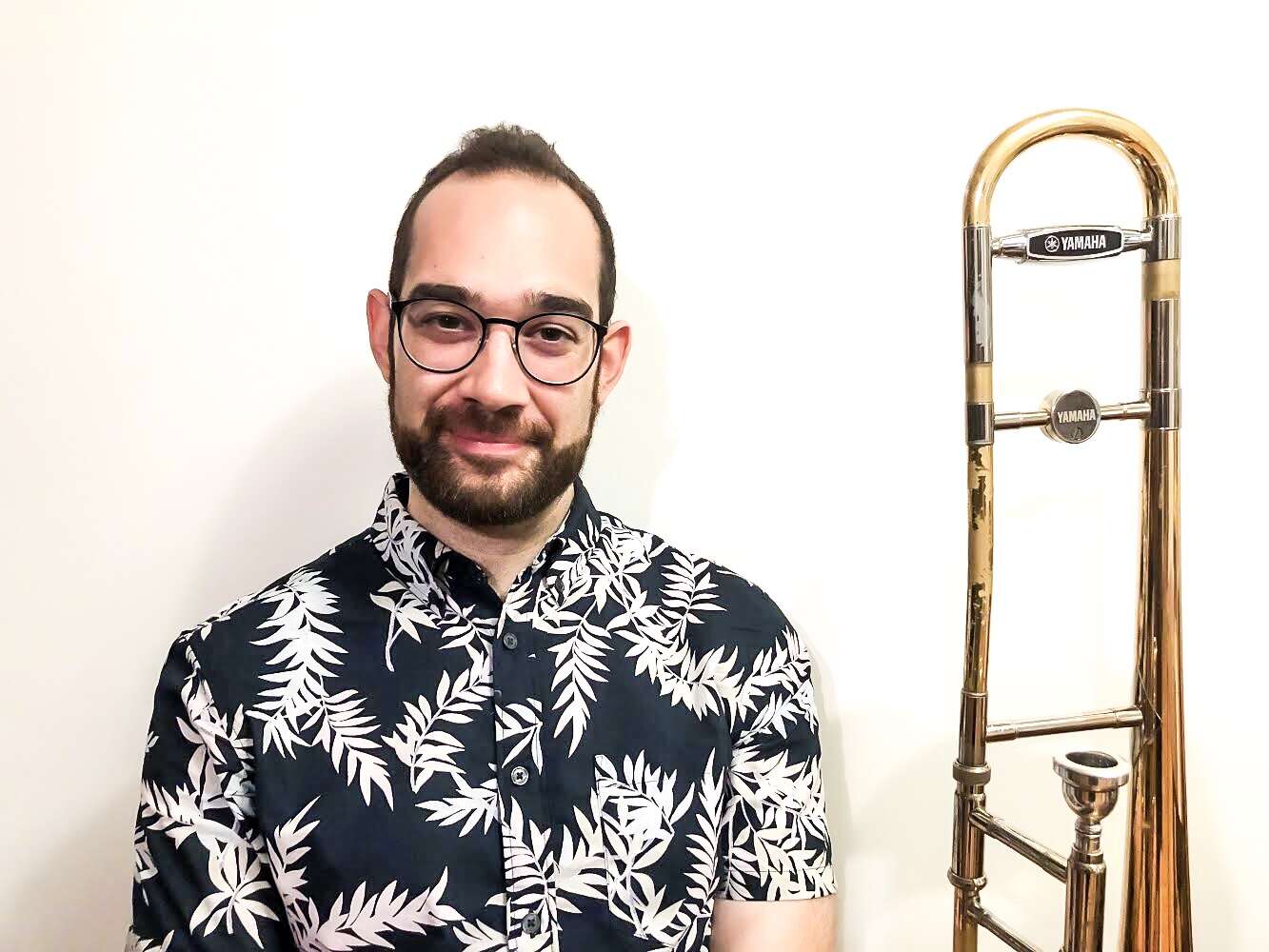 Person wearing a short-sleeve button-up shirt with a black-and-white leaf pattern, holding a trombone and smiling warmly.