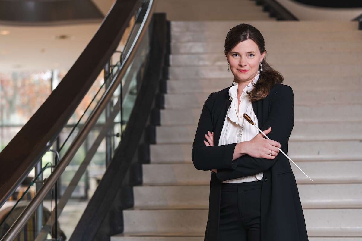 A person wearing a white blouse and black cardigan, standing on a white curved staircase with their arms crossed, holding a conductor's baton.