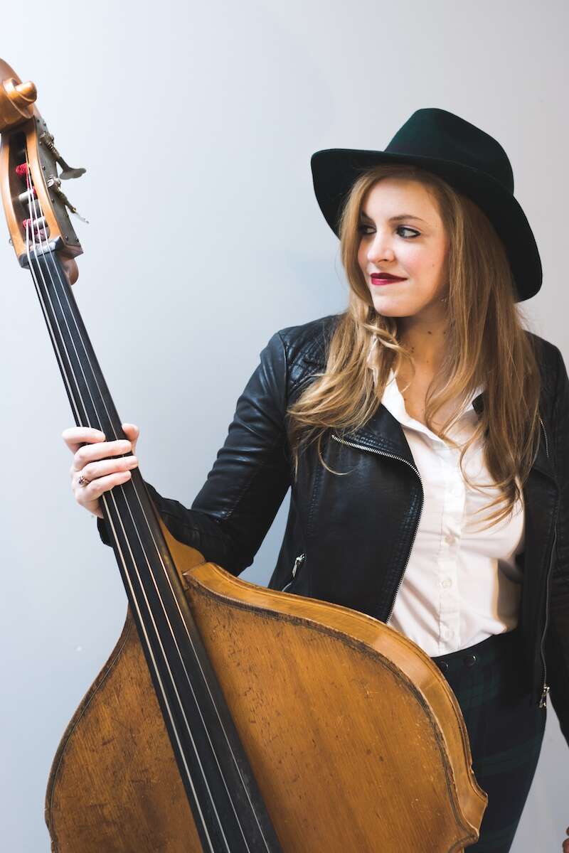 Person wearing a leather jacket and a cowboy hat, holding a double bass and looking to the side.