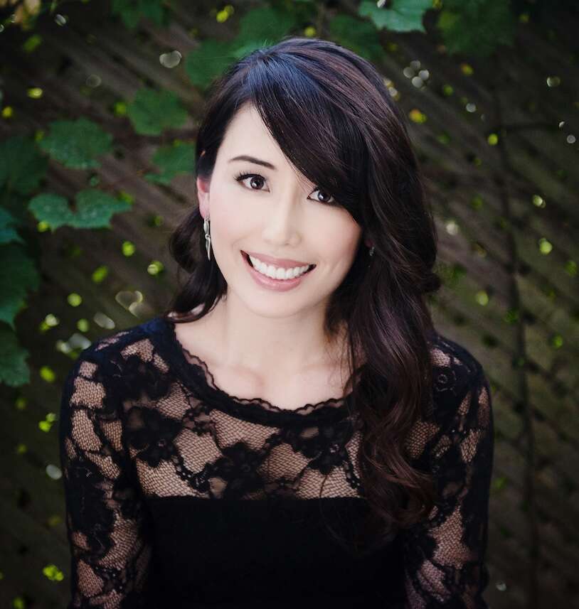 Person with long hair, wearing a black dress and smiling, standing in front of a lattice panel with leaves, creating a natural and elegant atmosphere.