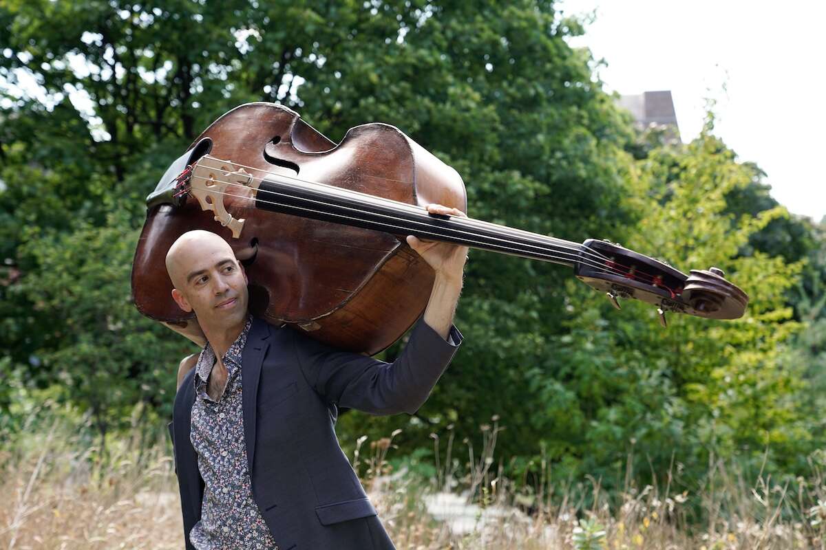 Person wearing a suit, holding a double bass behind their head in a field with trees in the background.