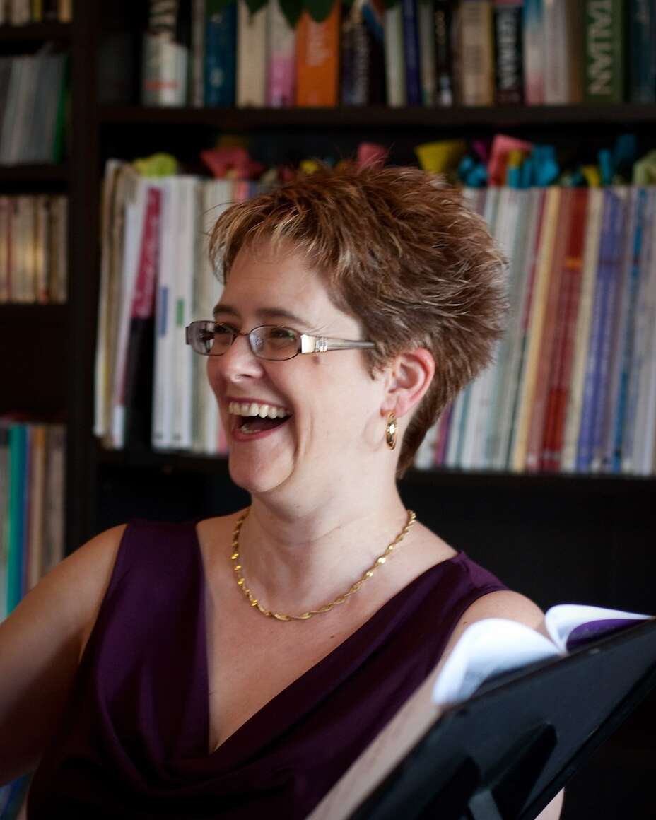 Person with short hair, wearing a purple dress, laughing while holding a book in a library.