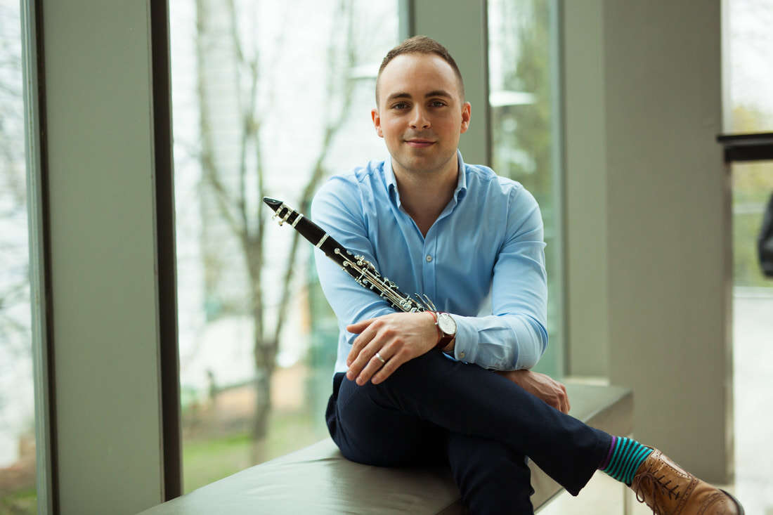 Person sitting down in front of a wall of windows, wearing business clothes and holding a clarinet.