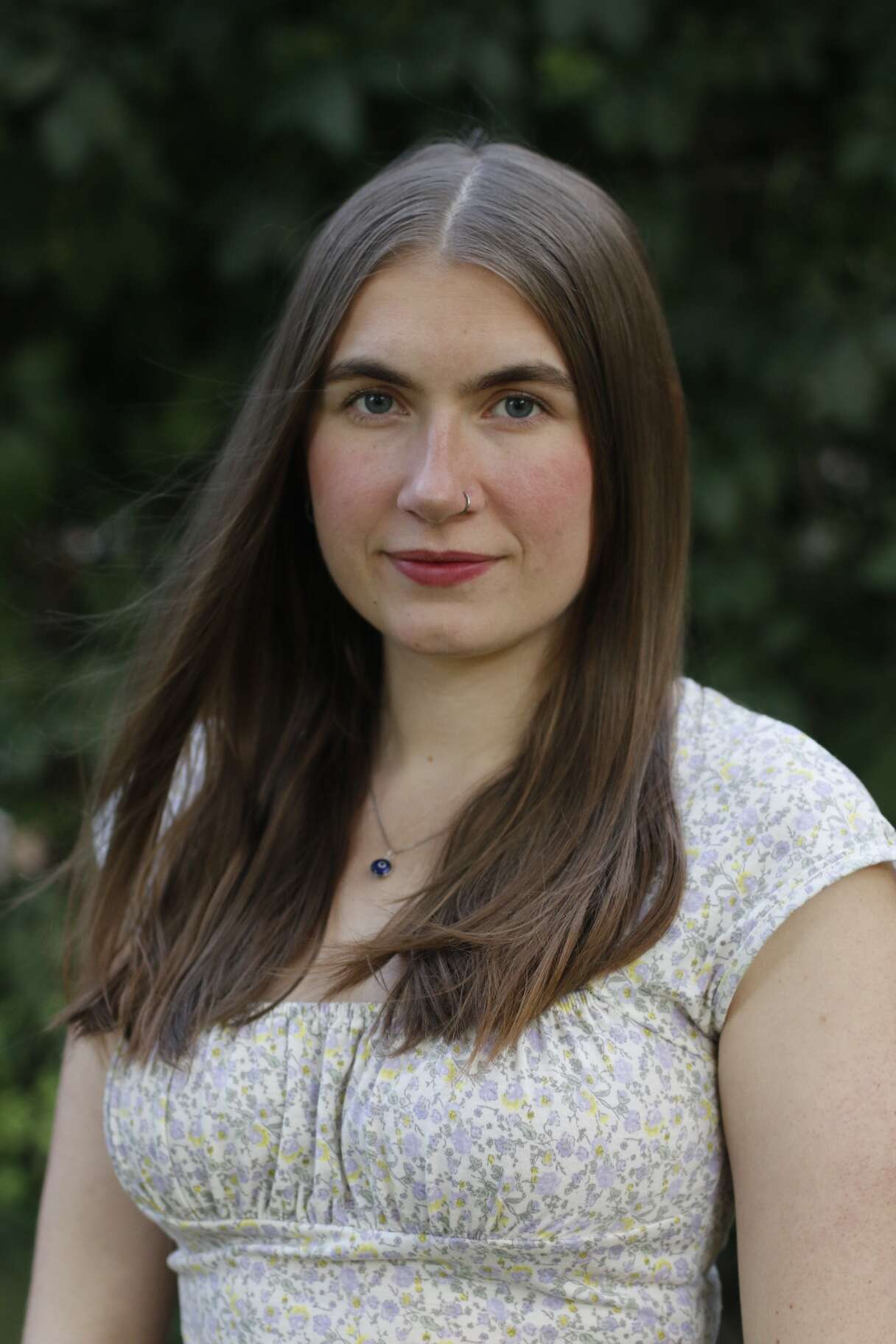 Young person with long hair wearing a white top. Blurred greenery in the background