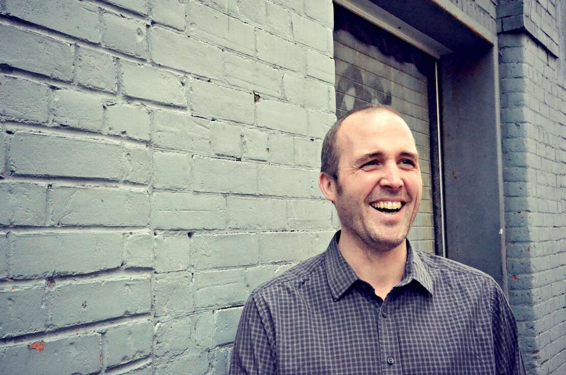 Headshot of a person smiling joyfully, wearing a grey button-down shirt, standing in front of a grey painted brick wall.