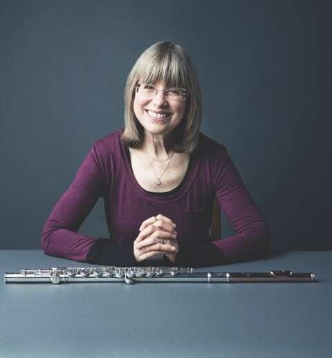 Person wearing a purple shirt, sitting at a table with their hands interlaced and a flute placed in front of them, smiling warmly against a dark table and background.