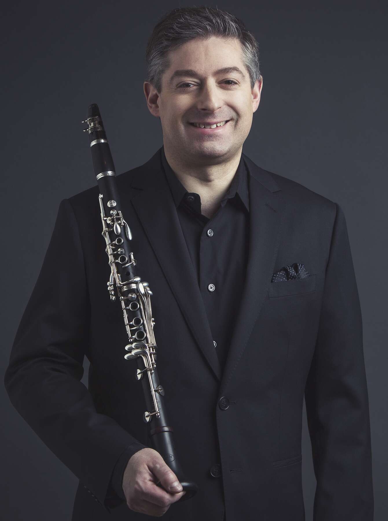 Person wearing a black suit, holding a clarinet and smiling, standing in front of a dark background, exuding sophistication and warmth.