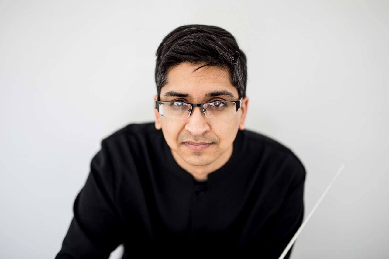 Headshot of a person wearing glasses and a black sweater, leaning slightly and holding a conductor's wand, with a light grey background.