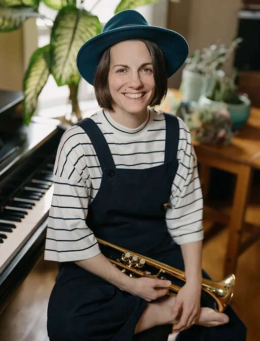Person wearing a blue hat and black dress, holding a trumpet while sitting on a piano stool in front of a piano, with plants surrounding the scene, creating a serene and artistic atmosphere.