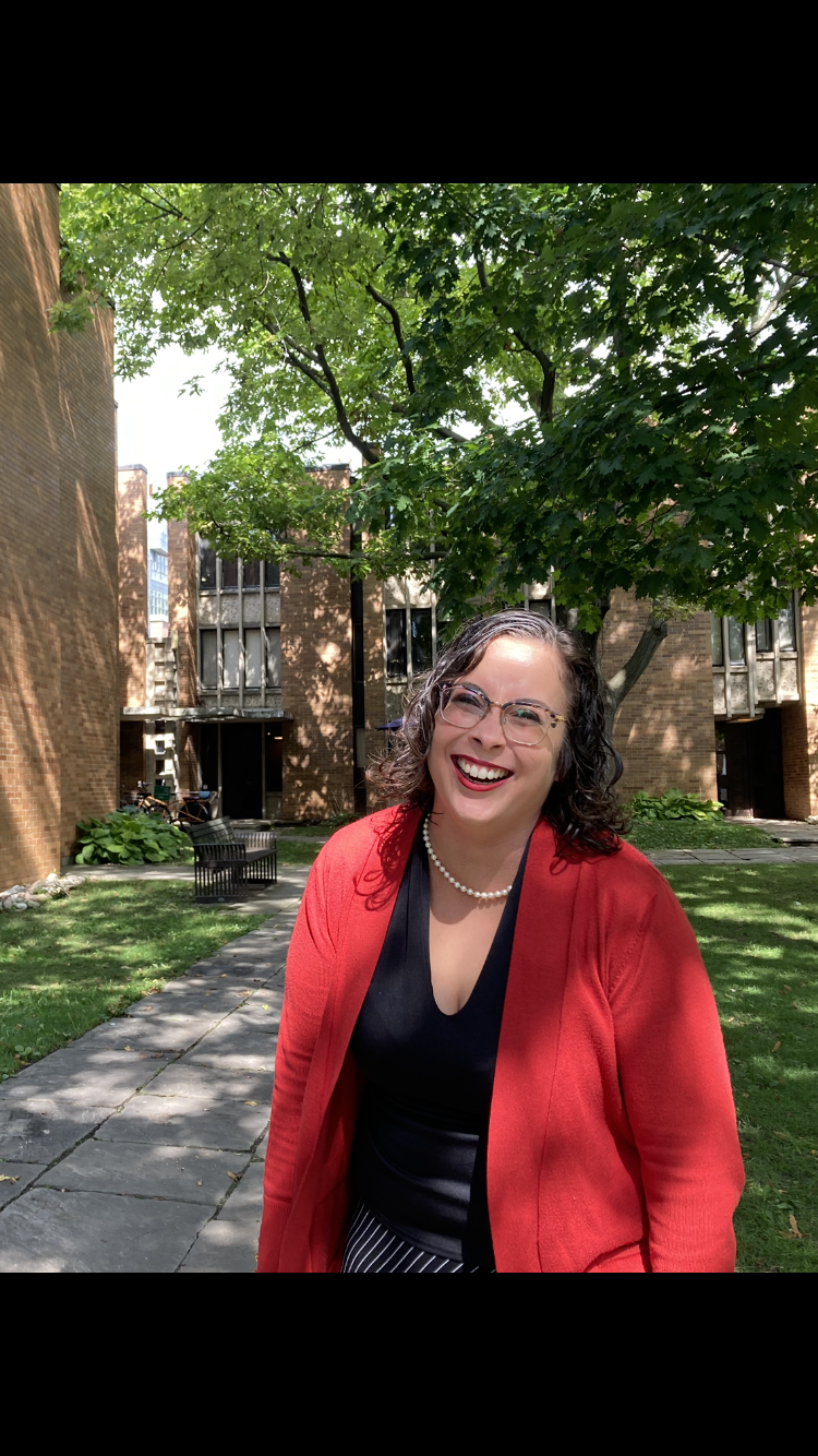 Person wearing glasses and a red jacket smiling. Standing on grass in front of tree and building