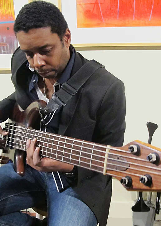 Person in a suit jacket and jeans playing an electric bass in front of a backdrop of colorful paintings.