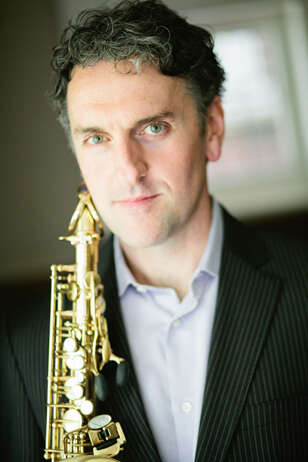 A headshot of a person wearing a white shirt and black suit jacket, holding a saxophone.