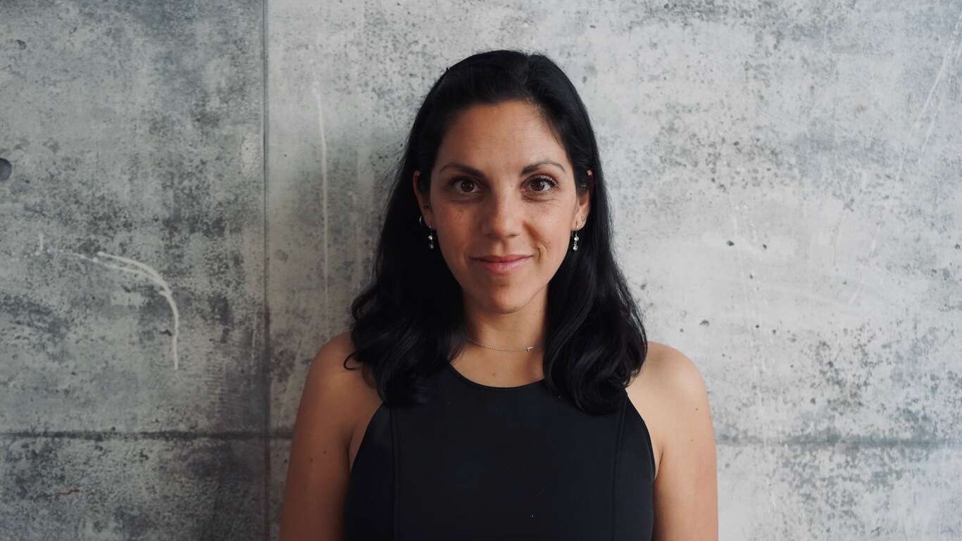 Person wearing a black dress, smiling, standing in front of a concrete wall.