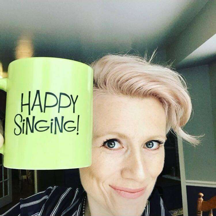 A person with short blonde hair grins while holding a green mug with the words 'happy singing' written on it, next to their face.