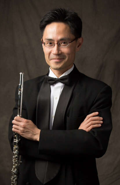 Person wearing a tuxedo, holding a flute in front of a brown background, presenting an elegant and refined appearance.