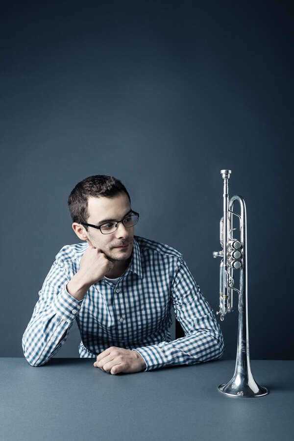 Person wearing a blue and white checkered shirt, with their hand on their chin, looking at a trumpet standing on the table. Both the background and the table are grey.