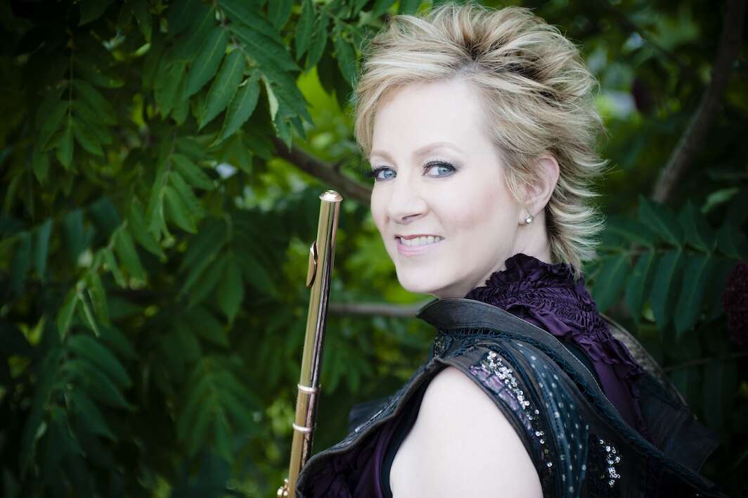 Headshot of a person looking over their shoulder, wearing black and holding a flute, with greenery in the background.