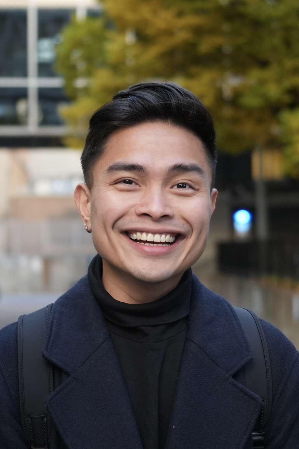 Headshot of a person wearing a black turtleneck and navy twill coat, smiling while standing in front of a tree and building.