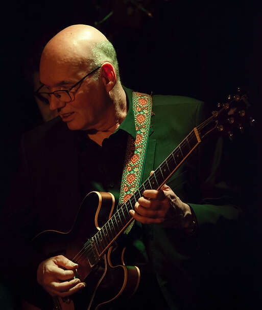 Person wearing all black playing the guitar, looking into the audience, illuminated by stage lights.