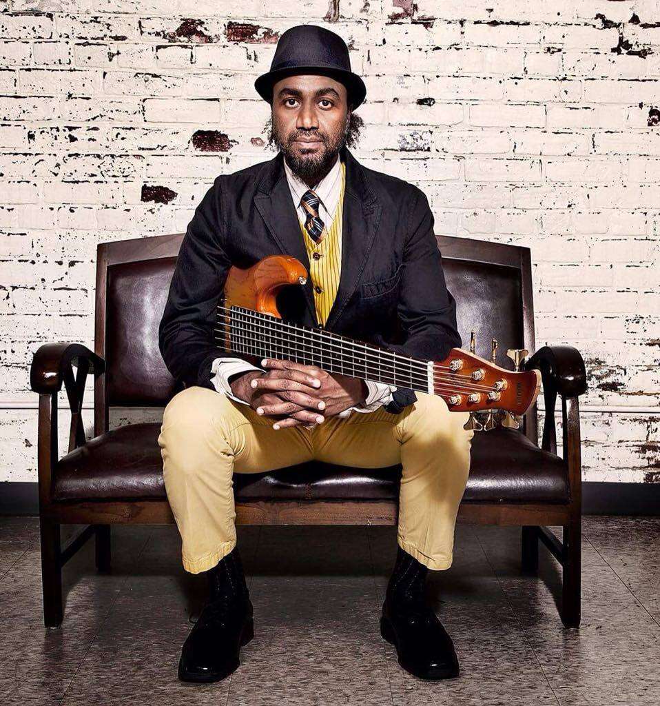 A person wearing a black and yellow suit with a black fedora, sitting on a bench with their fingers interlaced, holding a guitar in front of a white painted brick wall.