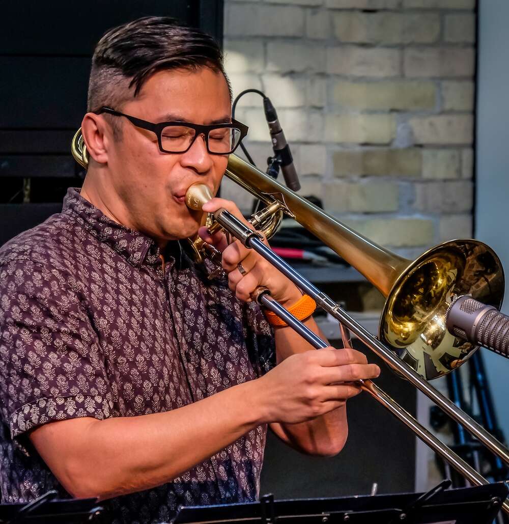 A person with glasses, wearing a red patterned short-sleeve button-up shirt, is playing the trombone while surrounded by sound equipment.