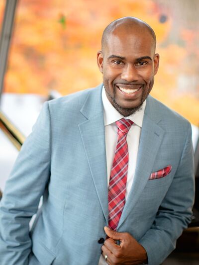 Portrait of a man smiling in front of a piano next to a window, wearing a grey suit with a red plaid tie.