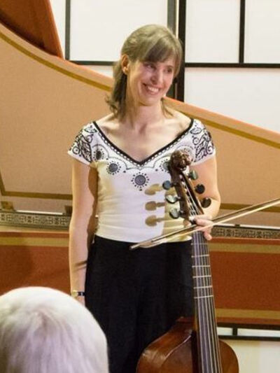 Picture of a woman standing in front of a piano, smiling and looking to the side while holding her instrument, wearing a white top and black pants.