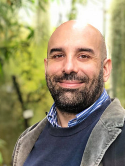 Portrait of a man with beard standing in a forest, surrounded by trees and natural greenery.