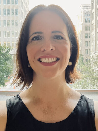 Headshot of a woman wearing a black shirt, smiling in front of a window with a city street in the background.