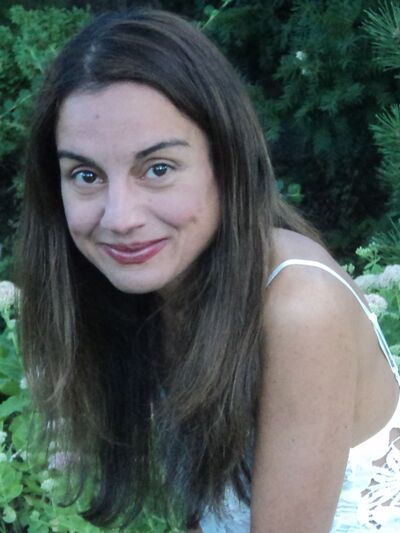Smiling woman with long hair wearing a white dress, sitting in a garden surrounded by plants and flowers.