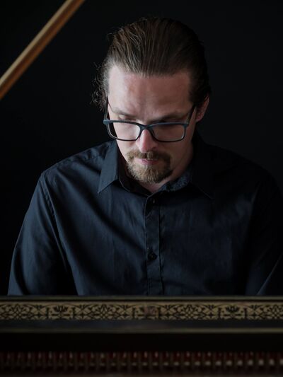 Person with longer hair wearing glasses and a black button-down shirt, playing the piano.