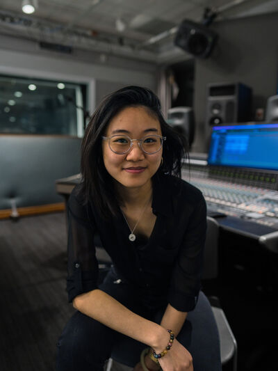 Person wearing a black button-down shirt sitting on a stool in the electronic music studio, surrounded by equipment and sound gear.